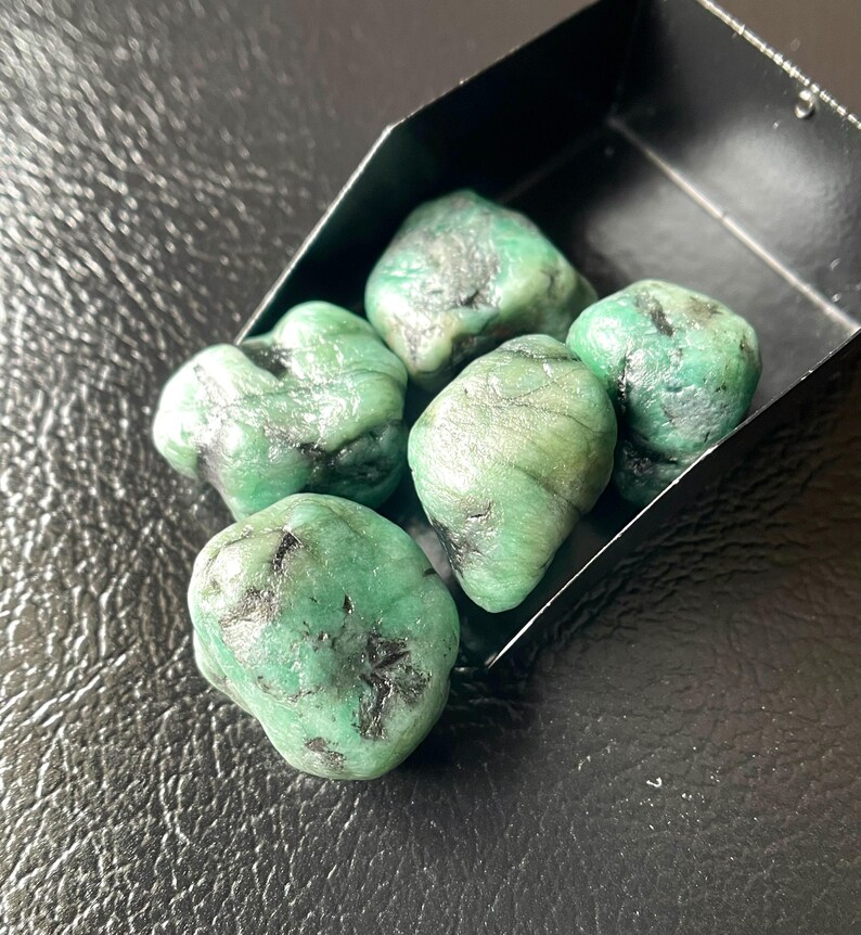 May include: A collection of raw emerald gemstones, each with a vibrant green hue and dark inclusions. The stones are displayed in a small, black container against a textured, dark surface. The emeralds vary in size and shape.