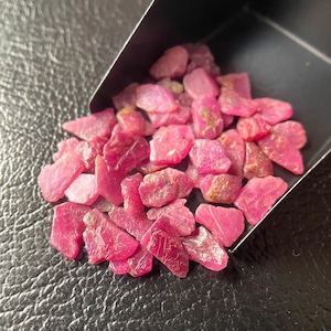 Op de afbeelding: Een verzameling ruwe, levendige roze robijn edelstenen. De edelstenen hebben verschillende vormen en maten, met een natuurlijke, ongepolijste uitstraling. Ze worden getoond in een kleine, zwarte container tegen een zwarte, getextureerde ondergrond.