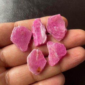 Op de afbeelding: Een verzameling van zes ruwe, ongeslepen robijn edelstenen. De robijnen hebben een levendige roze-rode kleur, met een enigszins doorschijnend uiterlijk. De stenen hebben onregelmatige vormen en zichtbare oppervlaktestructuren, wat wijst op een natuurlijke oorsprong.