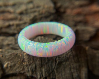 May include: A close-up of a white opal ring with iridescent colors. The ring is round and has a smooth, polished surface. The opal displays flashes of green, blue, and pink. The ring is set against a dark, textured background.
