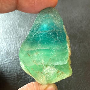 Puede incluir: Una gema en bruto, translúcida, con una mezcla de tonos verdes y turquesas. La piedra se sostiene entre los dedos, mostrando su textura natural e irregular y sus variaciones de color. Parece ser un espécimen mineral en bruto.