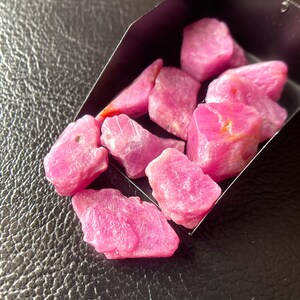Op de afbeelding: Een verzameling ruwe, roze robijn edelstenen, gepresenteerd in een kleine, donkere metalen container. De edelstenen hebben een natuurlijk, ongepolijst uiterlijk en tonen hun ruwe, kristallijne structuur. De achtergrond is een getextureerd, donker oppervlak.