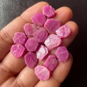 Op de afbeelding: Een verzameling ruwe, ongeslepen robijn edelstenen. De edelstenen hebben een levendige roze kleur, met een ruw, getextureerd oppervlak. De robijnen zijn onregelmatig van vorm en variëren in grootte. De edelstenen worden in een hand gehouden.