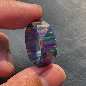 May include: A faceted, iridescent opal ring. The ring displays a spectrum of colors, including blues, greens, and reds, within its dark, translucent structure. The gemstone is held between fingers, showcasing its polished, geometric design.