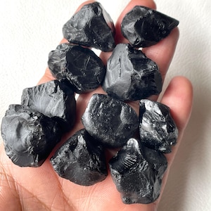 Puede incluir: Un conjunto de piedras de turmalina negra en bruto. Las piedras tienen forma irregular con un acabado mate y se sostienen en una mano sobre un fondo blanco. Las piedras varían en tamaño y tienen un aspecto natural y sin pulir.