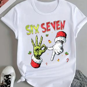Könnte beinhalten: Weißes T-Shirt mit einer Grafik, die zwei Hände zeigt, eine grüne und eine weiße, die die Zahl sieben bilden. Der Text "SIX SEVEN" steht in Grün und Rot über den Händen. Die Hände tragen Weihnachtsmützen und das Design enthält weihnachtliche Akzente.
