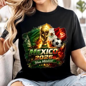 Puede incluir: Una camiseta negra de manga corta con un estampado gráfico del trofeo de la Copa Mundial de la FIFA, un balón de fútbol y la bandera de México. El diseño incluye el texto MEXICO 2026 y Viva México! con un efecto de fondo vibrante y ardiente.