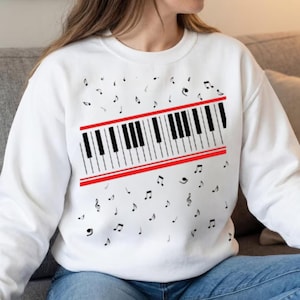 Pode incluir: Camisola branca com um design de teclado de piano em preto e branco, acentuado por uma faixa vermelha. Notas musicais dispersas circundam o gráfico. Uma peça de vestuário casual com tema musical.