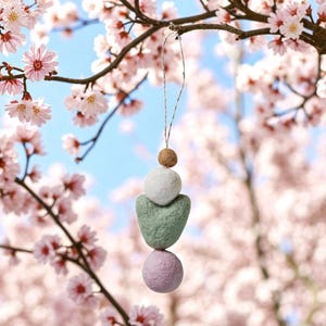 May include: A hanging ornament with a string, featuring a brown sphere, a white sphere, a green heart-shaped piece, and a lavender sphere. The ornament hangs from a tree branch with pink blossoms against a blue sky.