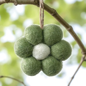 May include: A handcrafted felt flower decoration, featuring a white central ball surrounded by six green felt balls. It is suspended by a natural twine string. The ornament is designed to resemble a flower and hangs from a tree branch.
