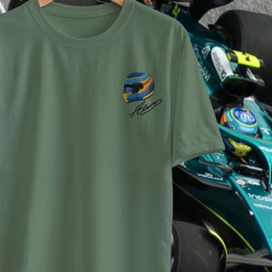 Op de afbeelding: Olijfgroen t-shirt met een kleurrijk racehelm ontwerp (blauw, geel, oranje) en een handtekening. Het t-shirt hangt aan een houten hanger. Op de achtergrond is een groene raceauto te zien.