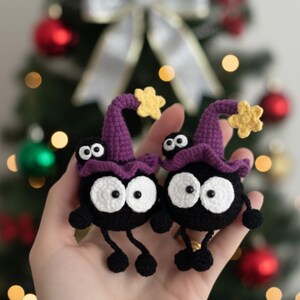 May include: Two black crochet figures with white eyes, wearing purple witch hats with yellow star accents. The figures have small black legs and arms. The background is a blurred Christmas tree with ornaments.