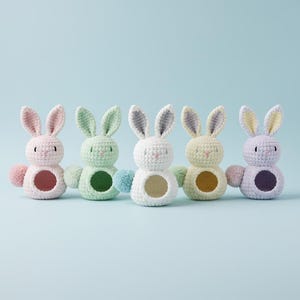 May include: Five pastel-colored crochet bunny figures with open centers and pom-pom tails. Each bunny has a different color scheme: pink, green, white, yellow, and lavender. The bunnies have long ears and are set against a light blue background.