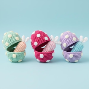 May include: Three crocheted Easter egg containers in pastel shades: mint green, hot pink, and lilac. Each egg is decorated with white spots and opens to show a crocheted carrot with white leafy tops. The eggs are set against a light blue backdrop.
