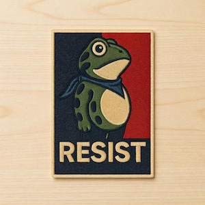 May include: Embroidered patch featuring a cartoon frog wearing a blue scarf, set against a red and navy blue background. The word "RESIST" is embroidered in tan letters at the bottom. The frog is green with dark green spots.