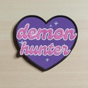May include: A heart-shaped patch with a purple background and the words "demon hunter" in pink script. The patch has a black border and small star accents.