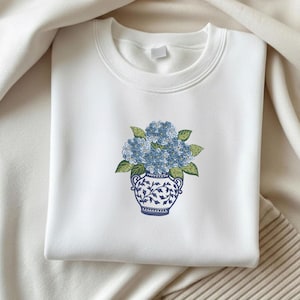 May include: White sweatshirt featuring an embroidered design of blue hydrangeas in a blue and white vase. The vase has a detailed floral pattern. The sweatshirt is made of a soft material and is suitable for casual wear.