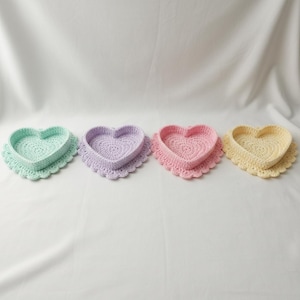 Könnte beinhalten: Vier herzförmige Häkeltabletts in Pastellfarben: Mintgrün, Lavendel, Rosa und Gelb. Jedes Tablett hat einen gewellten Rand und ist vor einem weißen Hintergrund platziert. Diese dekorativen Tabletts sind ideal für kleine Gegenstände.