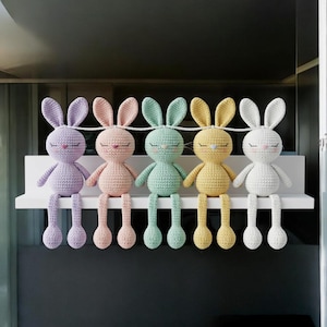 Könnte beinhalten: Fünf gehäkelte Hasenpuppen in Pastellfarben: Lavendel, Rosa, Mintgrün, Gelb und Weiß. Jeder Hase hat lange Ohren, geschlossene Augen und lange, herabhängende Beine. Sie sitzen auf einem weißen Regal vor einem dunklen Hintergrund.