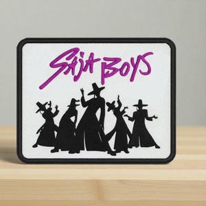 May include: Embroidered patch featuring the text "Saja Boys" in purple script above a silhouette of six figures in hats and long coats. The patch has a black border and is on a white background.