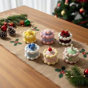 Puede incluir: Una colección de siete mini pasteles de ganchillo en varios colores, incluyendo amarillo, morado, azul y verde, cada uno con adornos decorativos únicos. Los pasteles están dispuestos sobre un camino de mesa de arpillera con decoraciones navideñas.