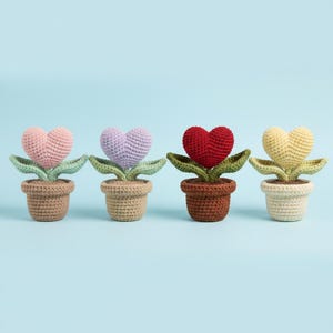 Peut inclure: Quatre fleurs en forme de cœur crochetées dans des pots. Les cœurs sont roses, violets, rouges et jaunes. Les pots sont marron et beige. Les feuilles sont vertes. Le fond est bleu clair.