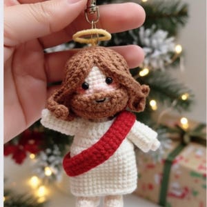 May include: Crocheted Jesus keychain with brown hair and beard, a red sash, and a gold halo. The figure is wearing a white robe. The keychain is held up against a blurred background of a Christmas tree and a wrapped gift.