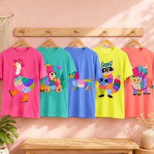 以下が含まれることがあります： 木製のラックに掛けられた5枚のカラフルなTシャツ。各シャツには、ガチョウ、クマ、ダックスフンド、アライグマ、牛など、ユニークなパッチワーク動物のデザインが施されています。シャツはピンク、ティール、ブルー、イエロー、コーラルの色合いです。