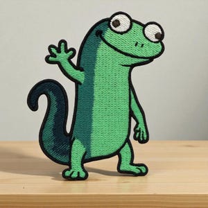 May include: Embroidered patch featuring a friendly green lizard with large, expressive eyes and a waving hand. The lizard has a dark green tail and is outlined in black. The patch is likely for clothing or accessories.
