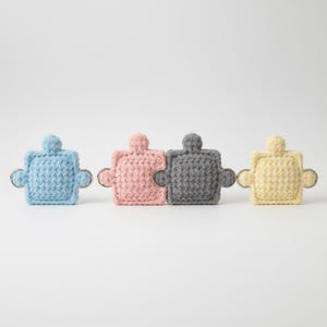 Puede incluir: Cuatro llaveros de piezas de rompecabezas de ganchillo en colores pastel: azul claro, rosa, gris y amarillo. Cada pieza tiene un pequeño lazo para sujetar las llaves. Los llaveros están dispuestos en fila sobre un fondo blanco.