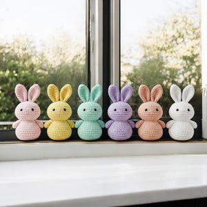 May include: Six pastel-coloured crochet bunnies sit on a windowsill. The bunnies are pink, yellow, mint green, purple, peach, and white. Each has long ears and a round body. Ideal for Easter decorations, these handmade bunnies are charming.