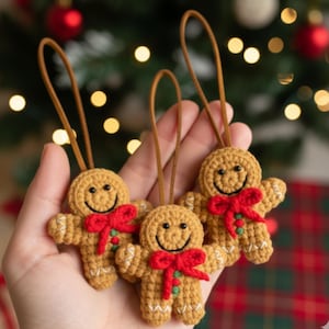 May include: Three crocheted gingerbread man decorations. Each has a brown body, white detailing, and a red bow. They feature smiling faces and brown hanging loops. The background shows a Christmas tree with festive lights and decorations.