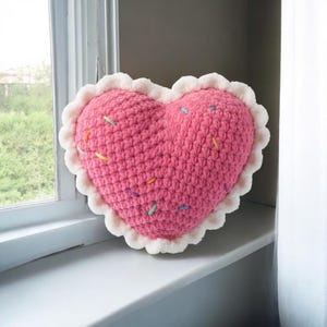 Puede incluir: Un cojín rosa en forma de corazón, tejido a ganchillo y adornado con chispas de colores. Tiene un borde blanco festoneado. El cojín está sobre un alféizar blanco.