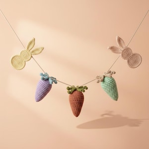Könnte beinhalten: Eine handgefertigte Häkel-Ostergirlande mit zwei Hasen-Silhouetten und drei Karotten in Lila, Orange und Grün. Die Girlande ist an einer dünnen, hellfarbigen Schnur aufgereiht, perfekt für Osterdekorationen.