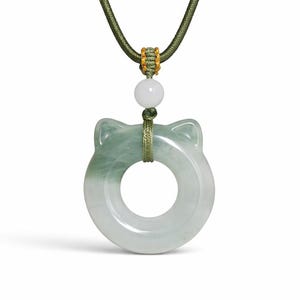 May include: A jade pendant necklace featuring a cat-shaped charm. The pendant is a light green color with a circular design and cat ear accents. The necklace has a green cord with gold accents and a white bead.