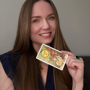 May include: A tarot card with a yellow sun design and the words "THE SUN" held up. The card is rectangular with a white border and is held by a person with long brown hair.
