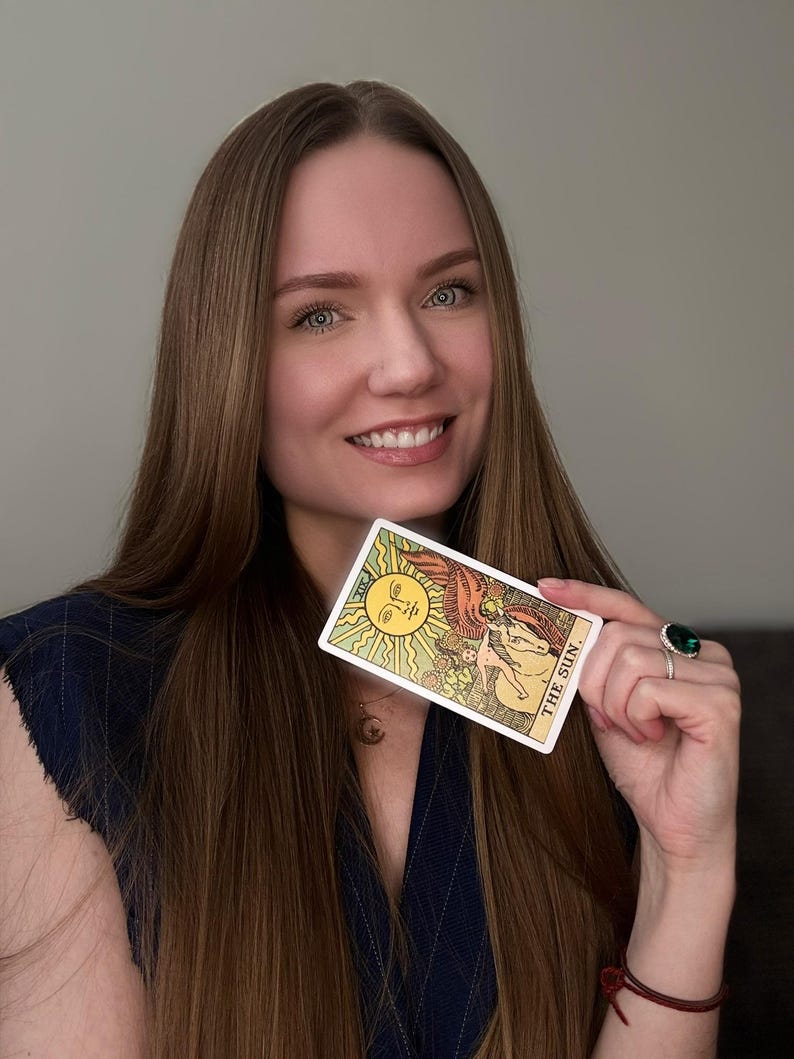 May include: A tarot card with a yellow sun design and the words "THE SUN" held up. The card is rectangular with a white border and is held by a person with long brown hair.