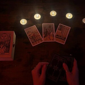 May include: A tarot card reading setup with lit candles and three face-up cards. A wooden box with a sun illustration and the text "THE SUN" is on the left. Hands hold a deck of cards.