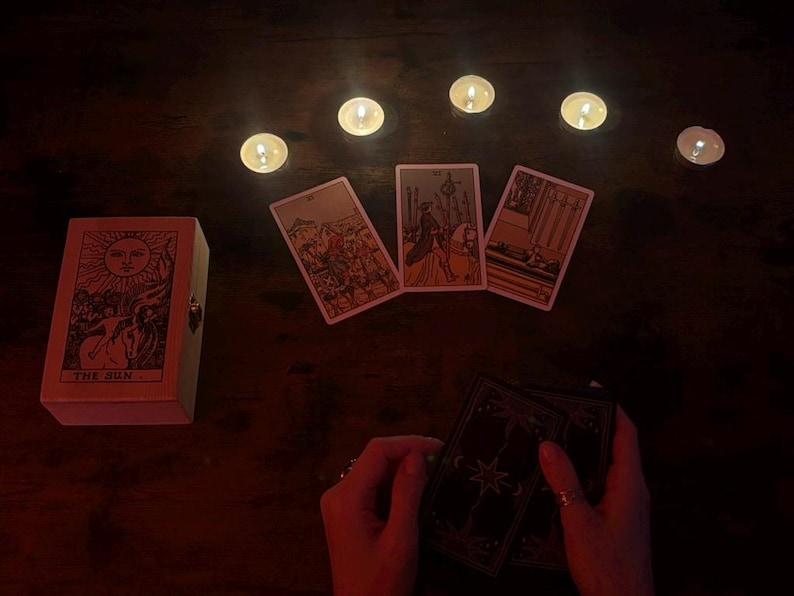 May include: A tarot card reading setup with lit candles and three face-up cards. A wooden box with a sun illustration and the text "THE SUN" is on the left. Hands hold a deck of cards.