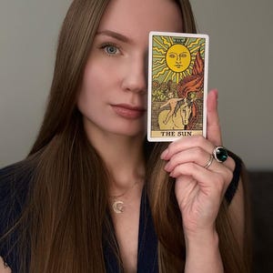 May include: A tarot card with a yellow sun, a white horse, and the text "THE SUN" is held up. The card is in front of a person's face. The person has long brown hair and is wearing a dark blue top.