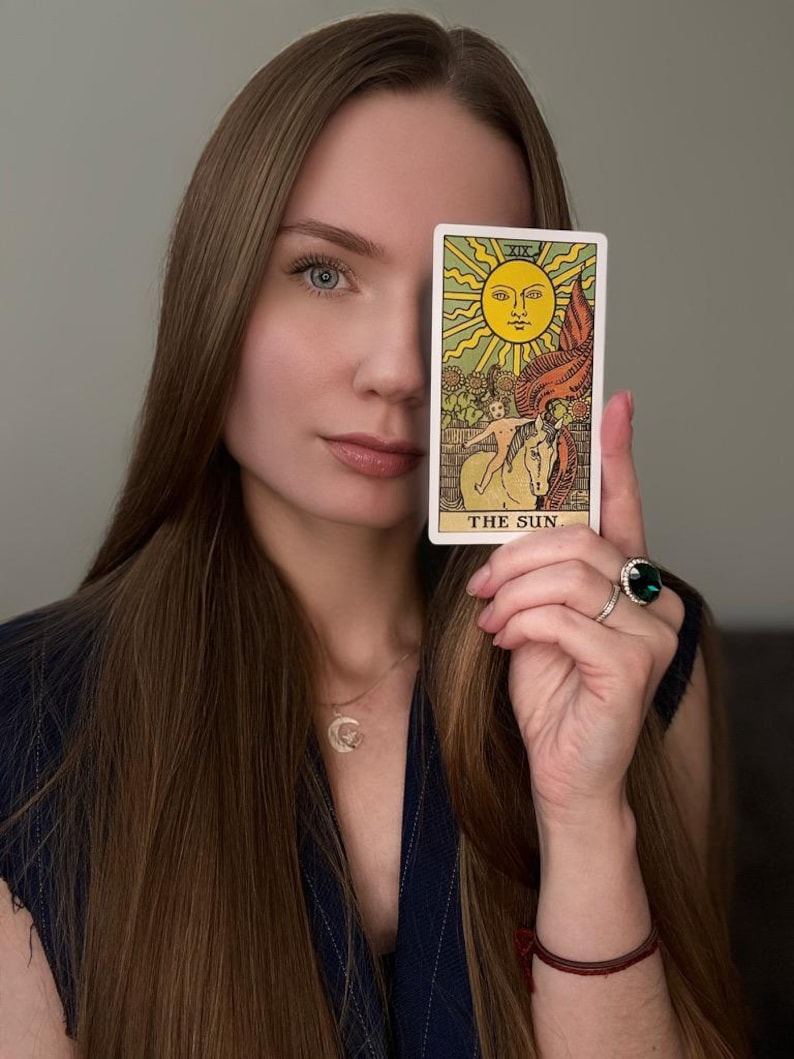 May include: A tarot card with a yellow sun, a white horse, and the text "THE SUN" is held up. The card is in front of a person's face. The person has long brown hair and is wearing a dark blue top.
