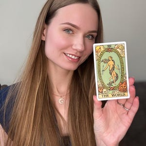 May include: A tarot card with the text "THE WORLD" held up. The card features a central figure within a laurel wreath, surrounded by figures and animals. The card has a white border and is held by a person.
