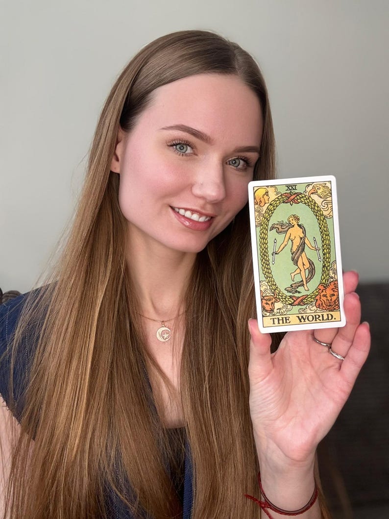 May include: A tarot card with the text "THE WORLD" held up. The card features a central figure within a laurel wreath, surrounded by figures and animals. The card has a white border and is held by a person.