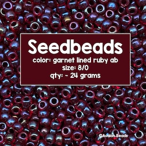 May include: A close-up of a pile of small, red, iridescent seed beads. The beads are 8/0 size and weigh approximately 24 grams. The text "Seedbeads" is written in white on a red background, along with the details "color: garnet lined ruby ab", "size: 8/0", and "qty: ~ 24 grams". The text "Avalon Beads" is written in white on a red background in the bottom right corner of the image.