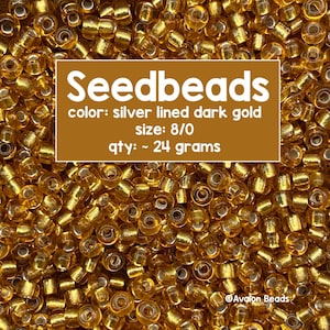 May include: A close-up of a pile of small, round, gold seed beads with a silver lining. The beads are approximately 8/0 size and weigh about 24 grams. The text "Seedbeads" is written in a brown rectangle above the beads, along with the details "color: silver lined dark gold", "size: 8/0", and "qty: ~ 24 grams". The text "Avalon Beads" is written in white at the bottom right corner of the image.
