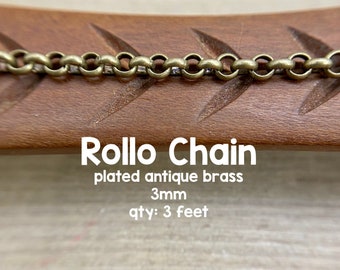 May include: A close-up of a 3mm antique brass plated rollo chain. The chain is 3 feet long and is shown on a wooden surface.  The text "Rollo Chain" is written in white on a brown background. The text "plated antique brass" is written in white on a brown background. The text "3mm" is written in white on a brown background. The text "qty: 3 feet" is written in white on a brown background. The text "Avalon Beads" is written in white on a brown background.