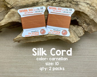 Puede incluir: Dos carretes de cordón de seda natural de color carneola, de 2 metros de largo y 0,90 mm de grosor, para enhebrar perlas y cuentas. Los carretes están etiquetados con "Natural Silk 100% Naturseide 2 m No. 10 0,90 mm" y "For stringing pearls and beads www.griffin.de". El texto "Silk Cord color: carnelian size: 10 qty: 2 packs Avalon Beads" está impreso debajo de los carretes.