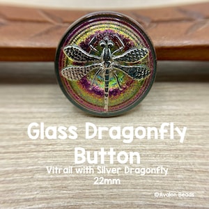 May include: A 22mm glass button with a silver dragonfly design. The button has a stained glass look with iridescent colors of purple, green, and gold.