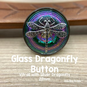 May include: A round glass button with a silver dragonfly design. The button has a vibrant, iridescent finish with green, purple, and blue hues. The button is 22mm in diameter.