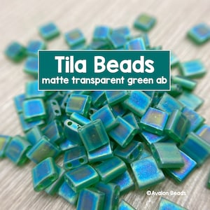 Può includere: Un mucchio di piccole perline quadrate di tila opache di colore verde con un bagliore iridescente. Le perline sono traslucide e hanno due fori. Il testo "Tila Beads matte transparent green ab" è visibile nell'immagine.